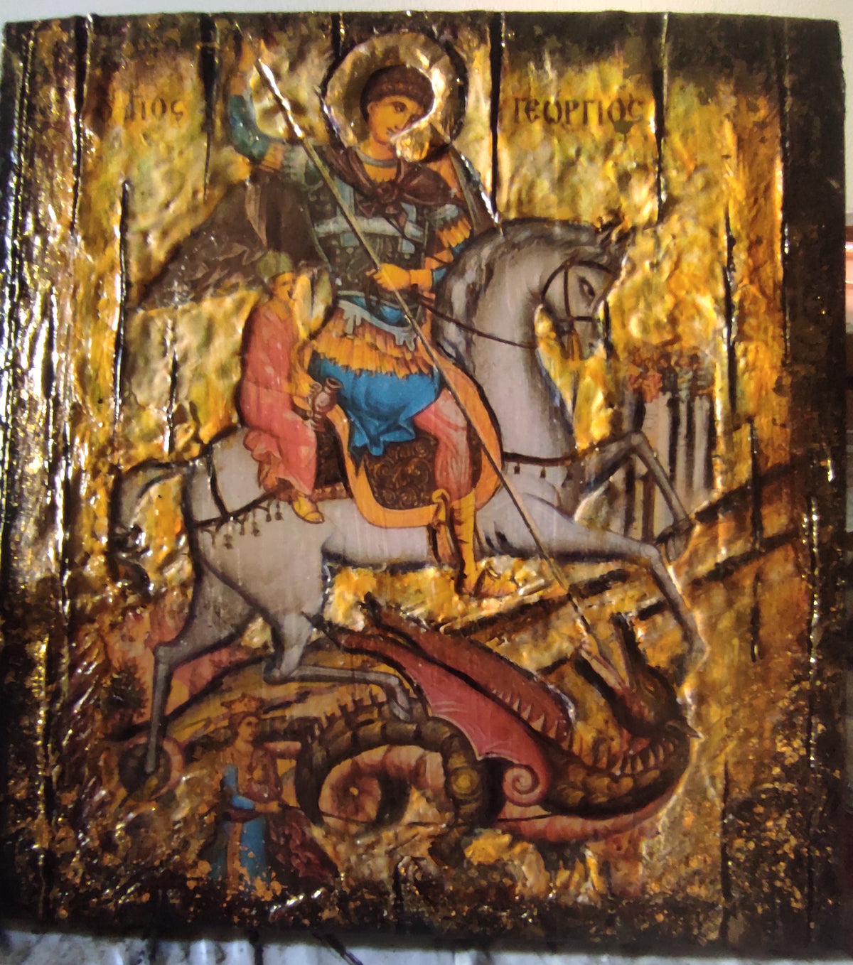 A Byzantine-style religious icon design of Saint George on a white horse, rendered with gold leaf and dark brown tones, photographed against a light fabric background.