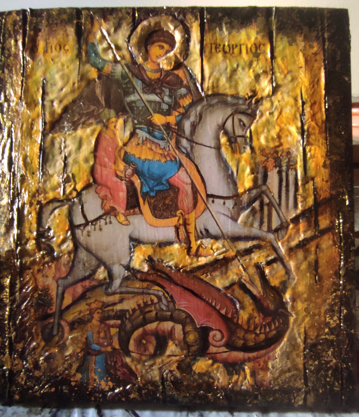 A rectangular religious icon design  on a wooden panel, depicting Saint George on a white horse with a red dragon beneath. The artwork has a distressed, textured finish with gold leaf, dark brown wood, and design figures in blue, red, and green.