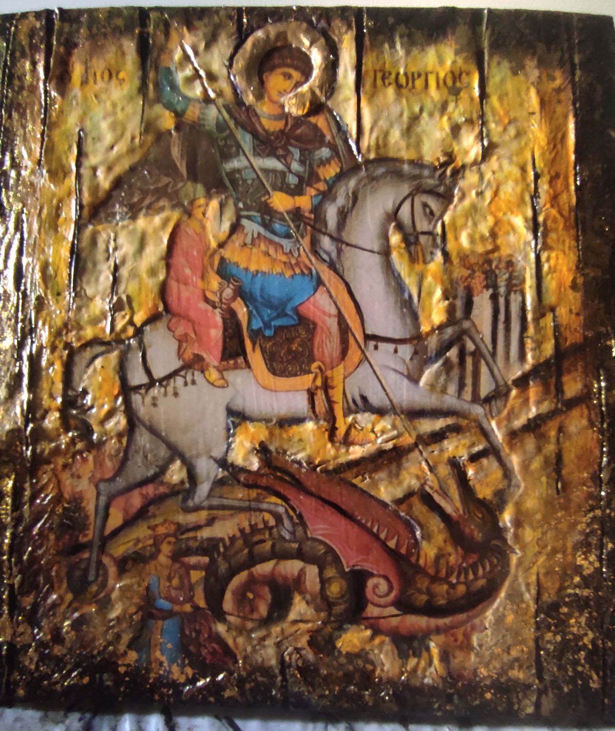 A Byzantine-style religious icon design on a gold leaf background, depicting Saint George on a white horse with a dragon beneath him, with a white cloth surface visible at the bottom.