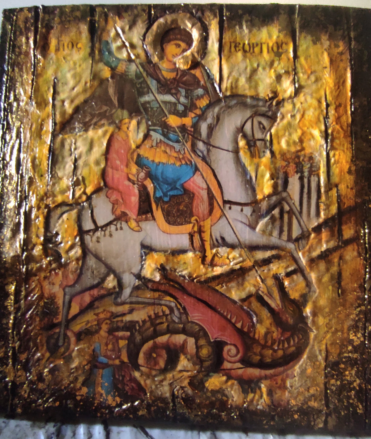 A Byzantine-style wooden icon design of Saint George on a white horse, with a gold leaf background and a red dragon beneath him, photographed flat against a light fabric surface.