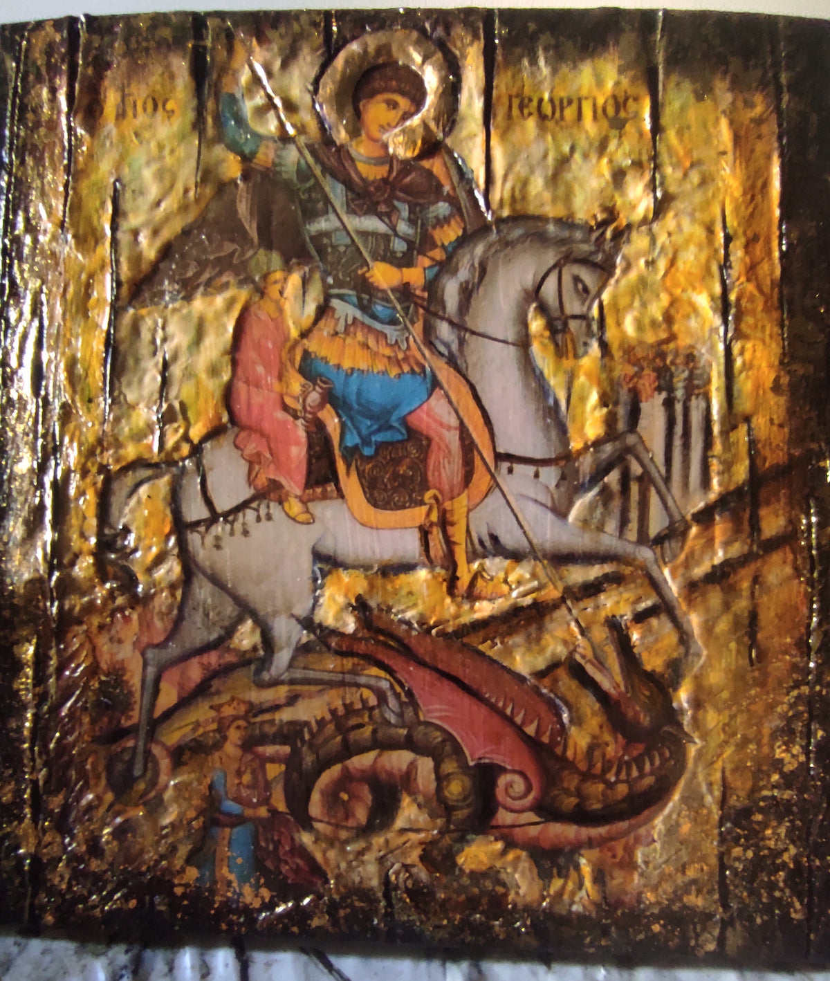 A Byzantine-style religious icon design of Saint George on a white horse, with a gold leaf background and dark brown distressed edges. The icon depicts Saint George in blue and red garments with a sword, standing atop a dragon.