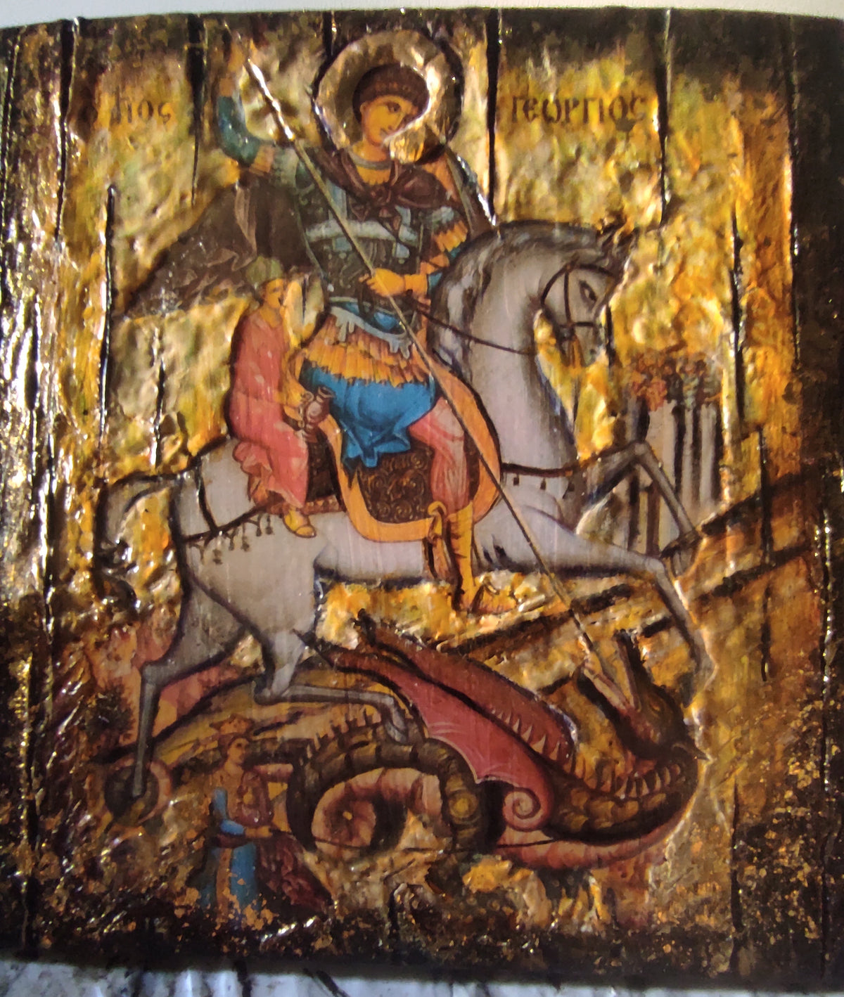 A rectangular religious icon design showing a saint on a white horse with a dragon beneath, rendered in metallic gold leaf with red, blue, and brown details. The artwork fills most of the frame, with a white cloth surface visible below it.
