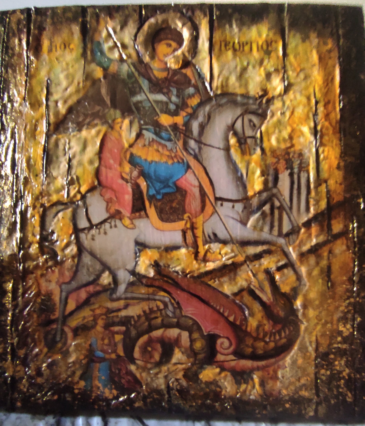 A square religious icon design on a gold leaf background depicting Saint George on a white horse, with a red dragon beneath him. The artwork has a textured, gilded appearance with dark brown and black accents.