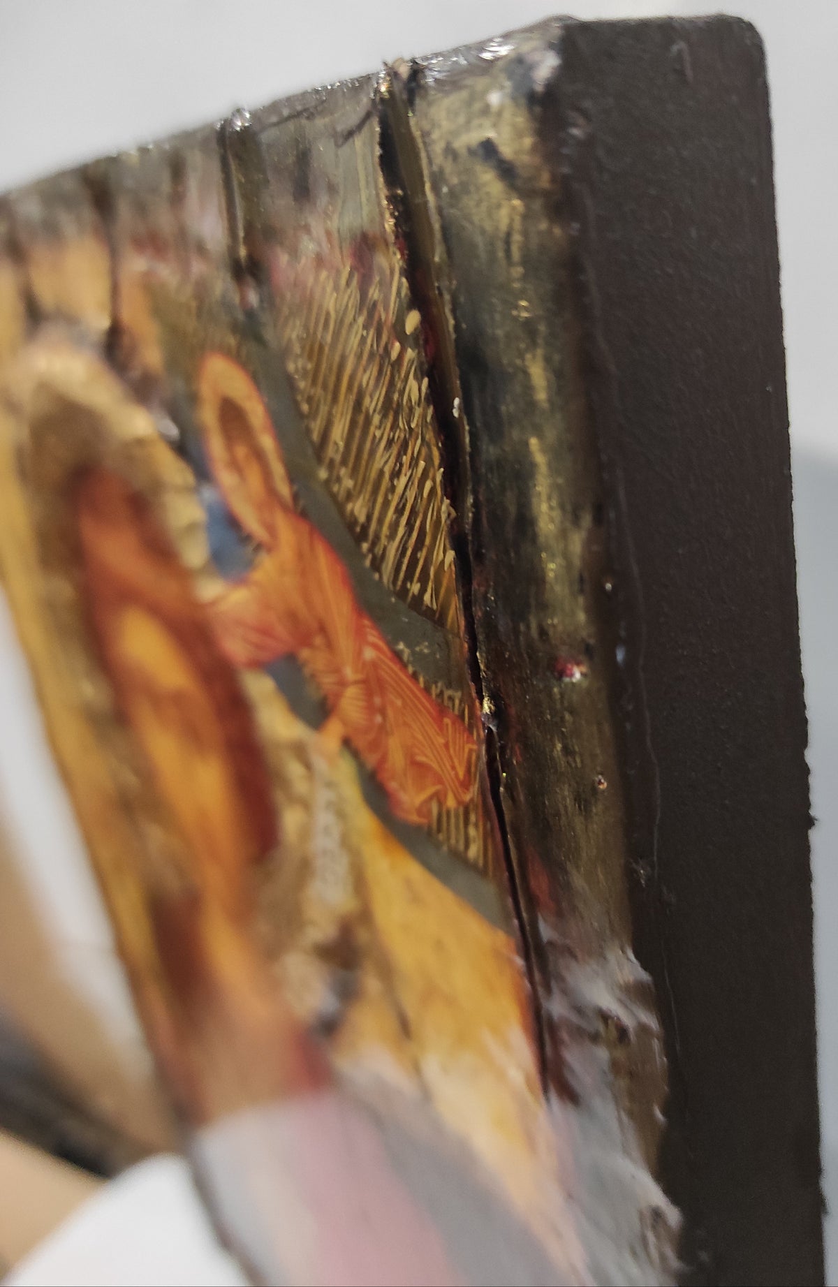 Close-up of a wooden religious icon panel showing the side edge with gesso and painted details of an angel and figure in warm gold, orange, and brown tones.