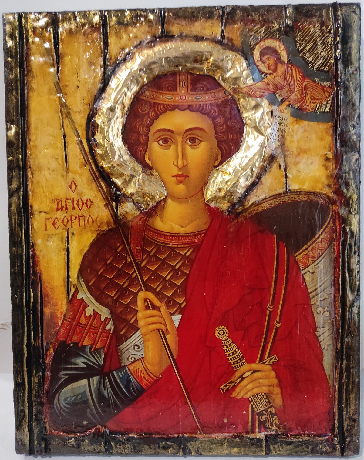 An antique wooden religious icon of Saint George on a white background. The icon features a gold leaf halo, a red and brown robe, and a sword, with a small angel in the upper right corner.