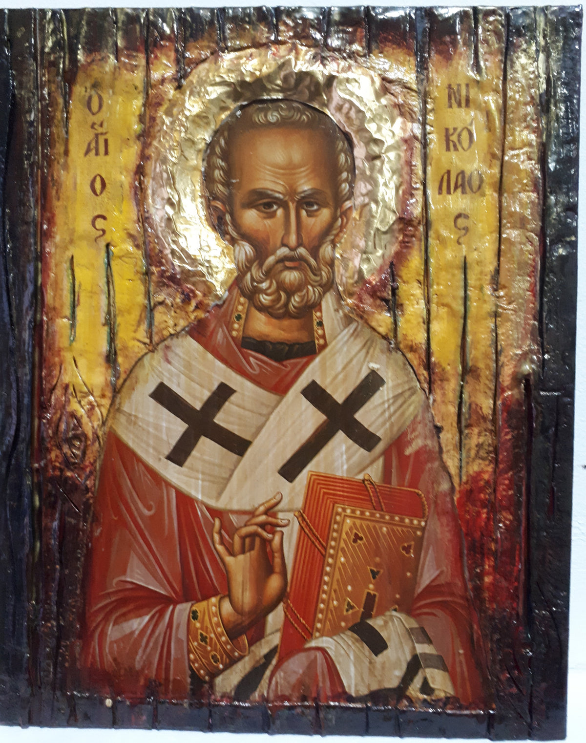 A rustic wooden religious icon design of Saint Nicholas, featuring a gold leaf halo, red and white robes with black crosses, and a book, mounted on a dark wooden panel.