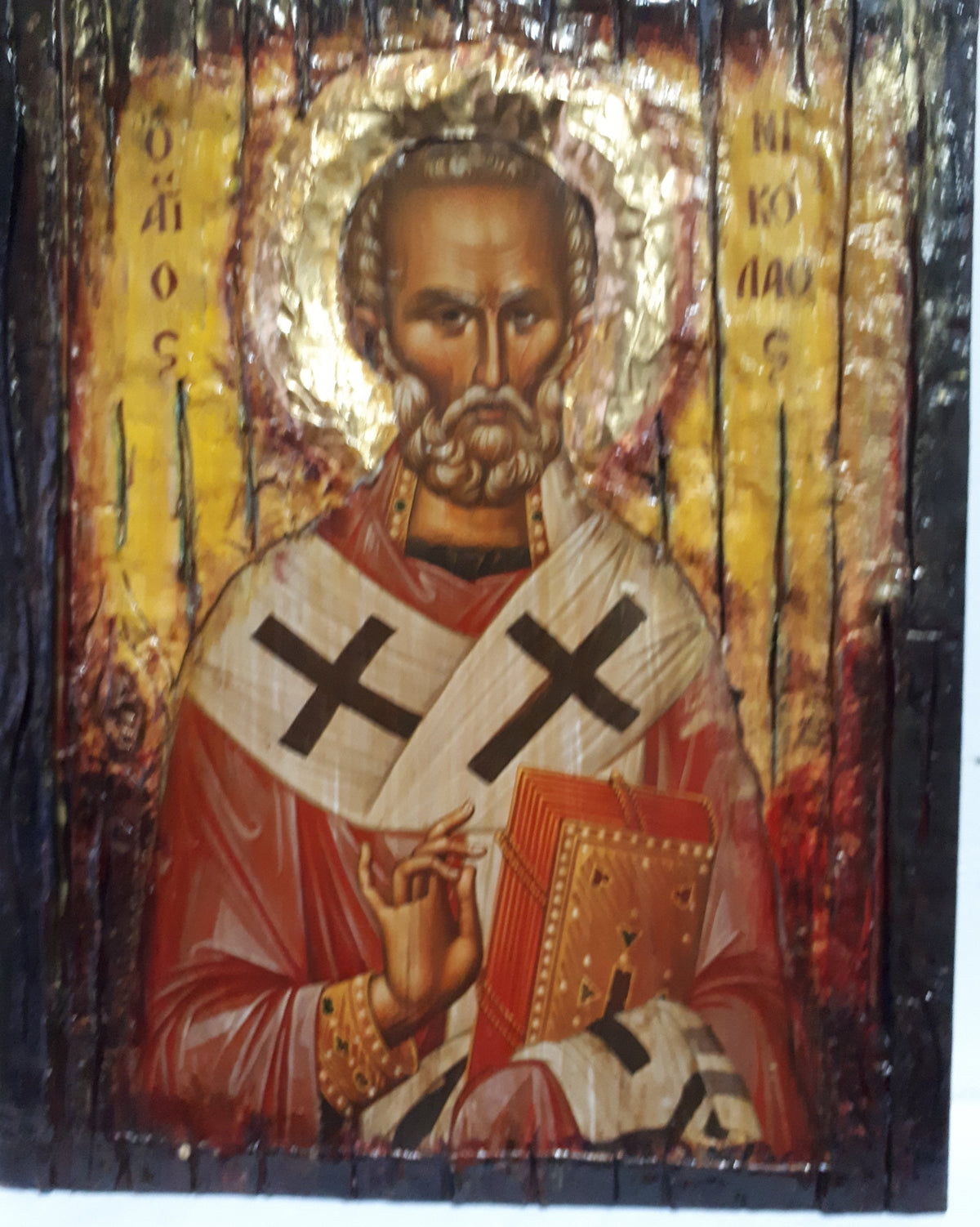 A rustic wooden religious icon design of Saint Nicholas, featuring a gold halo, red and white robes with black crosses, and a book, mounted on a dark wood panel.