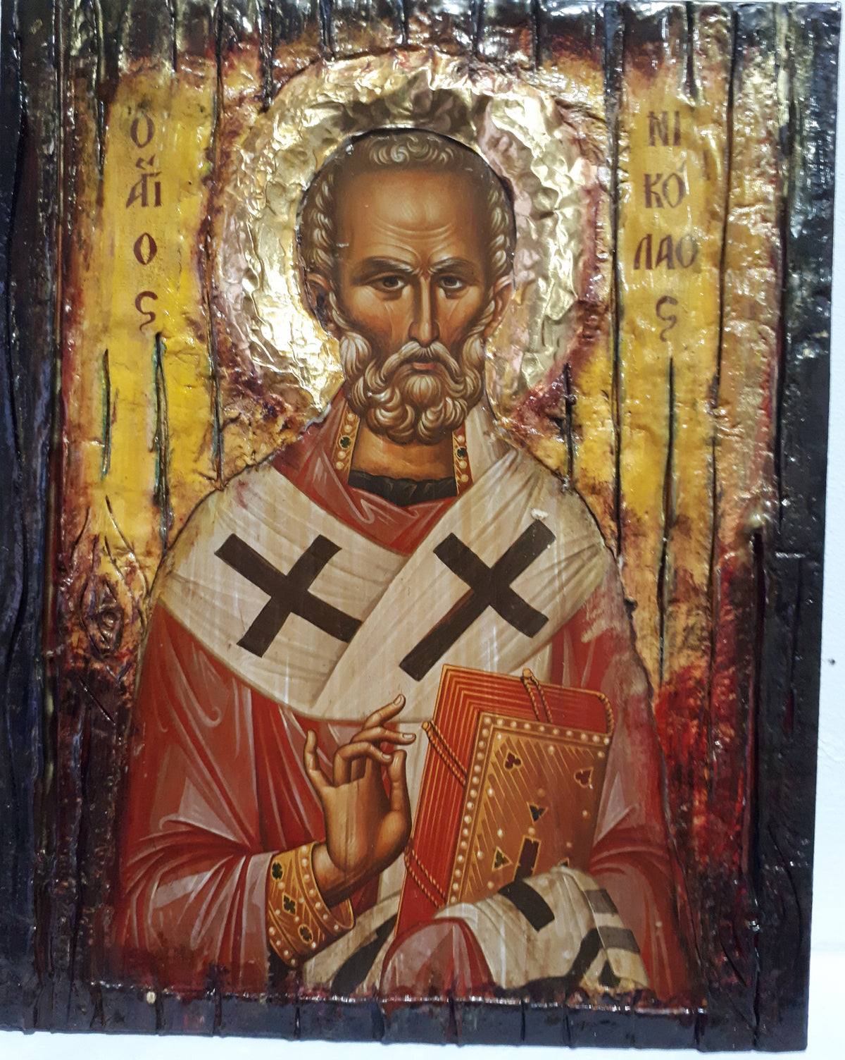 A rustic wooden religious icon of Saint Nicholas wearing a red and white robe with black crosses, holding a book, set against a gold background with Cyrillic inscriptions.