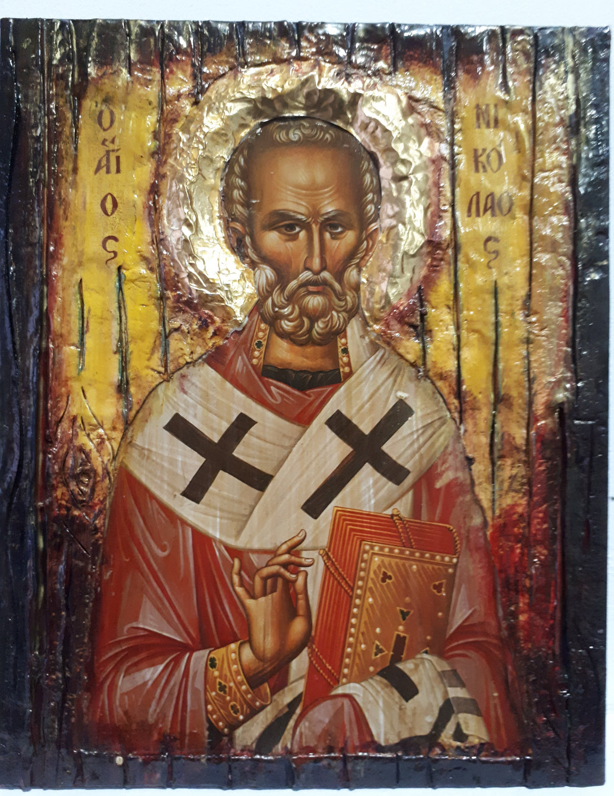 A rustic wooden religious icon of Saint Nikolas holding an open book, featuring a gold leaf halo and red and white robes on a distressed dark wood background.