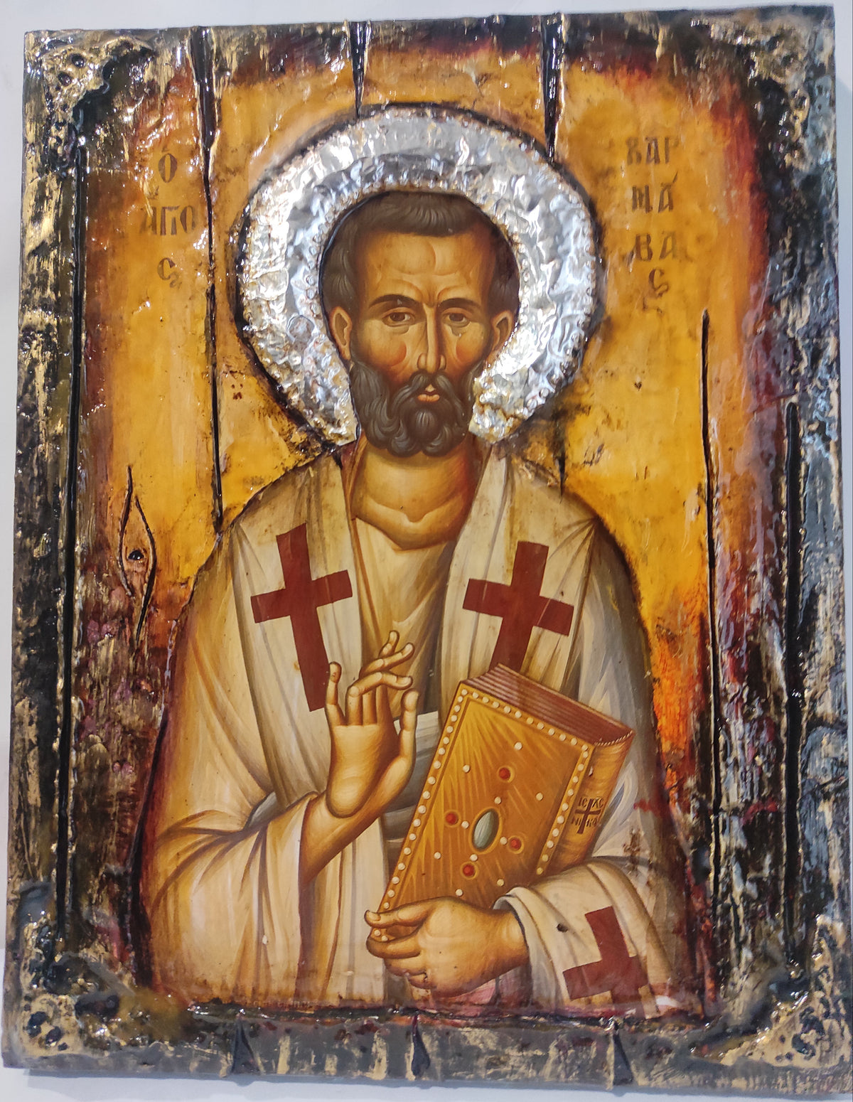 A rustic, hand-design Eastern Orthodox icon of Saint Barnavas  on a wooden panel. The icon features a textured gold background, a silver foil halo, and a distressed black and brown border, with Barnavas depicted in white robes holding a gold book and making a blessing gesture.