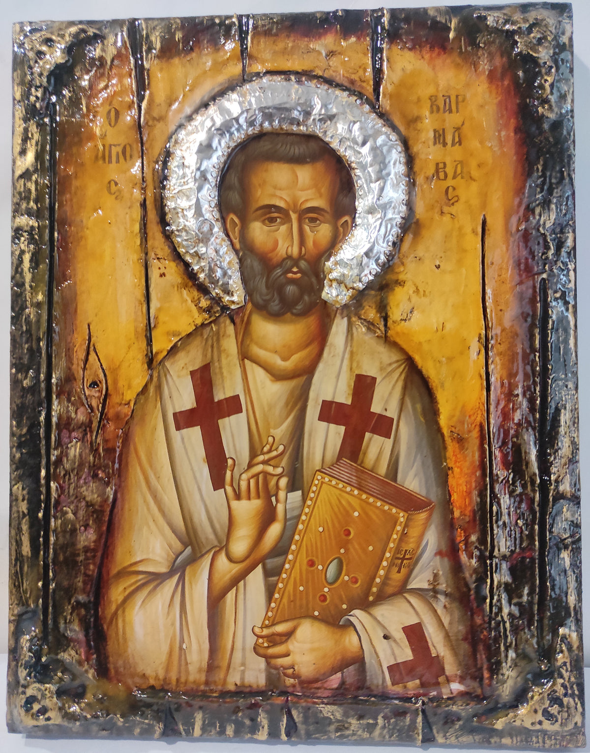 A rustic religious icon design of Saint Varnavas on a distressed wooden panel. The saint is depicted with a silver foil halo, wearing a white robe with red crosses, and holding a gold book.