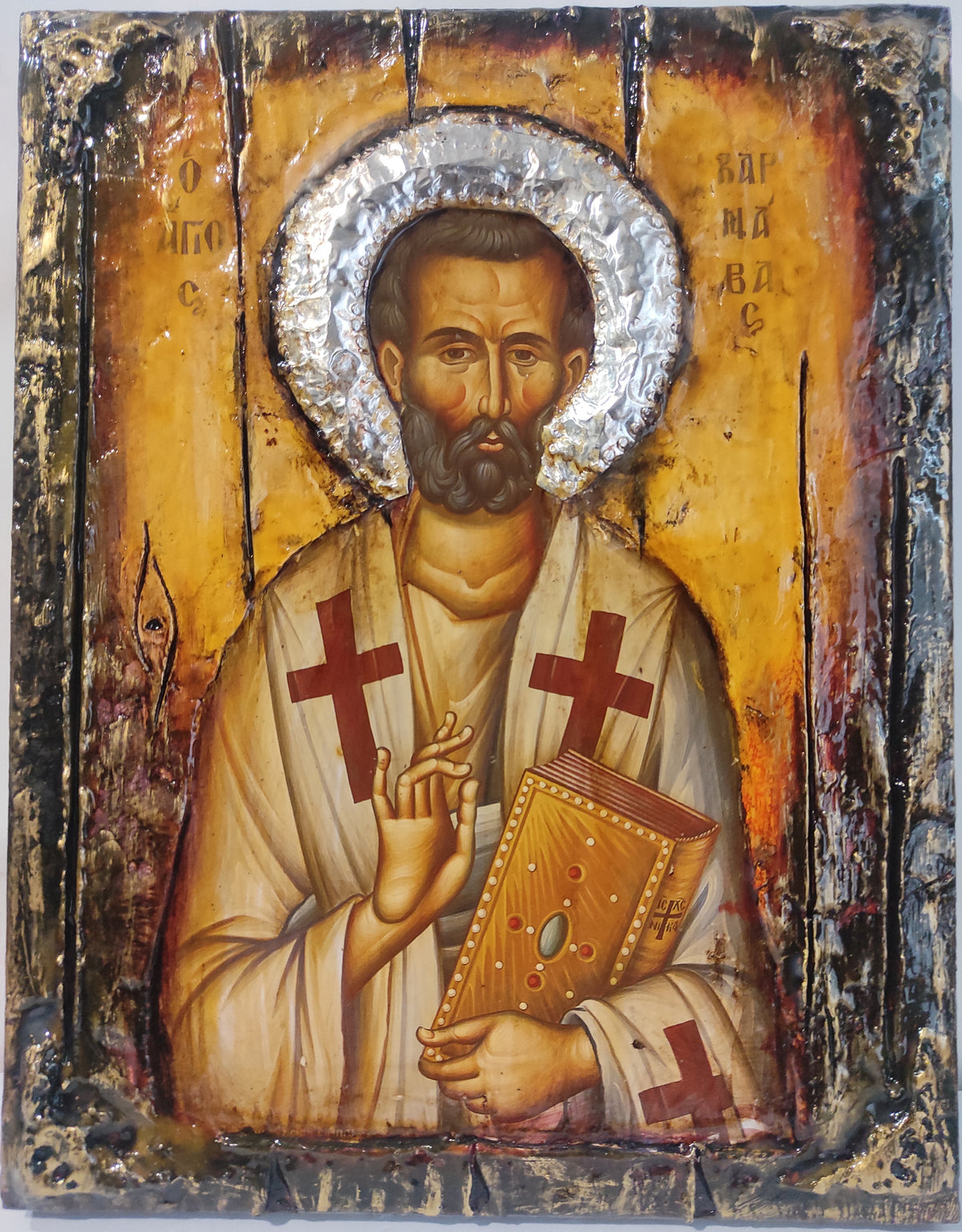 A rustic religious icon design of Saint Barnavas, featuring a metallic silver foil halo, a gold background with distressed black and brown textures, and the saint in white robes holding a book with red crosses.