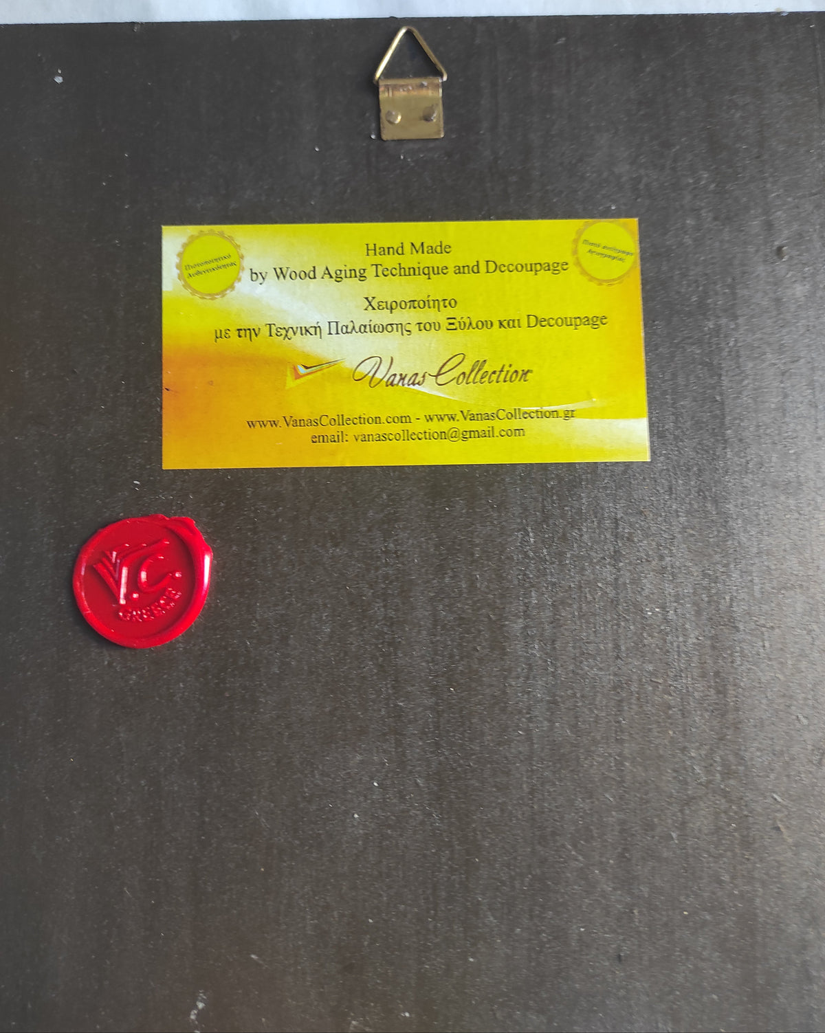 A square dark gray wood panel with a brass hanging loop at the top, a yellow rectangular label in the center reading 'VanasCollection' and 'Hand Made by Wood Aging Technique and Decoupage', and a red wax seal at the bottom left.