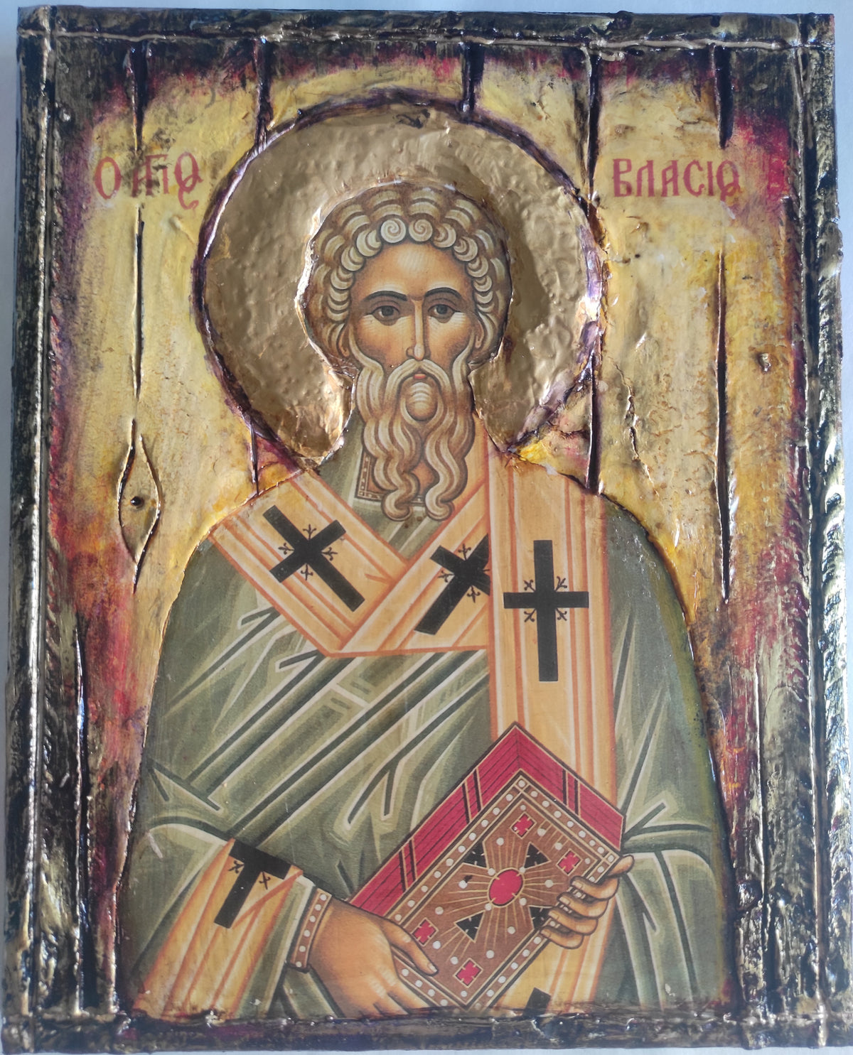 A religious icon painting of Saint Vlasios on a textured wooden panel with gold leaf and dark metal borders. The saint is depicted with a halo, wearing green and gold robes with black crosses, holding a book, with red lettering at the top.