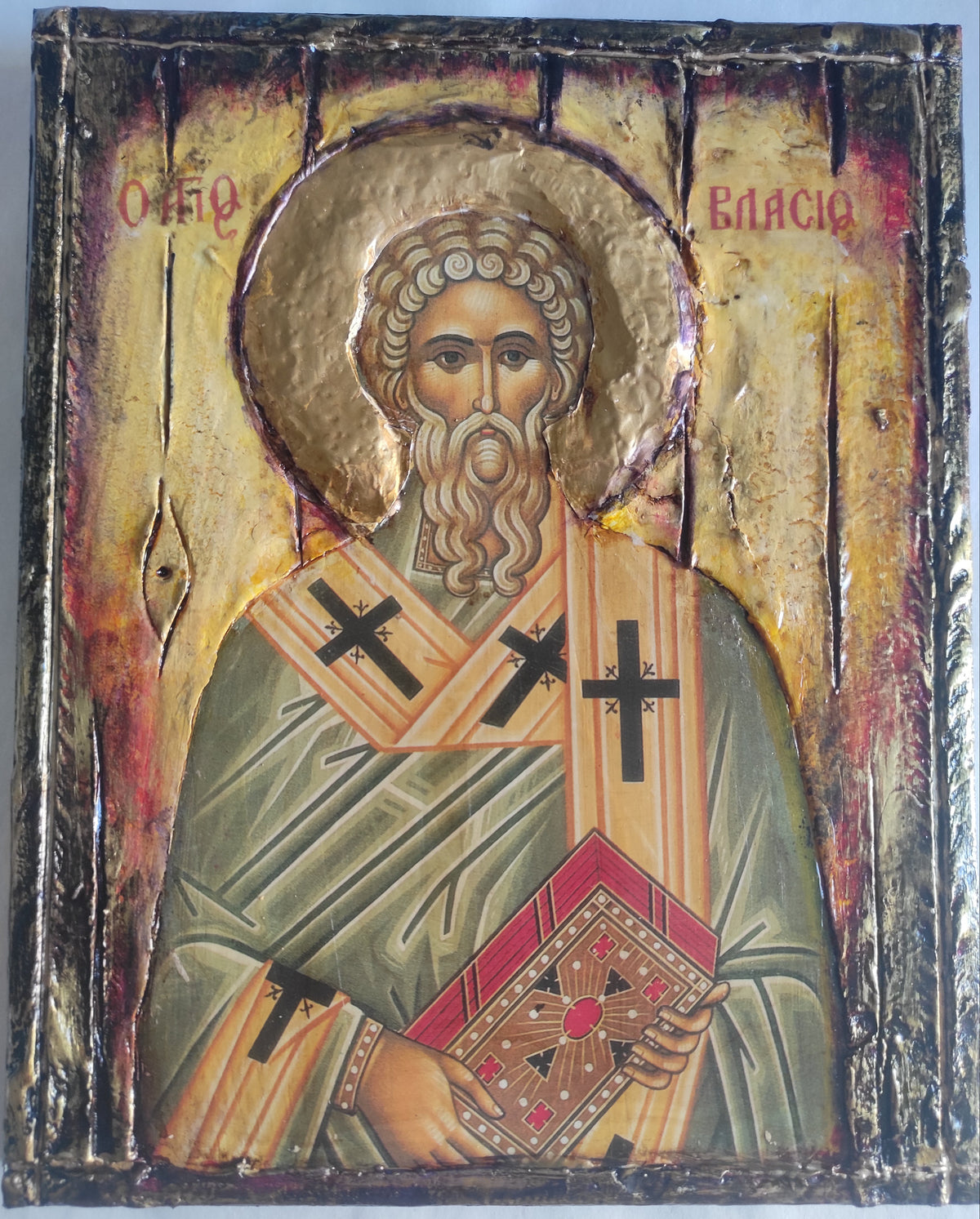A rectangular religious icon design on a metallic gold background, depicting a saint in green and gold robes with red accents and a halo. The icon has a distressed, textured finish with dark borders and Cyrillic lettering at the top.