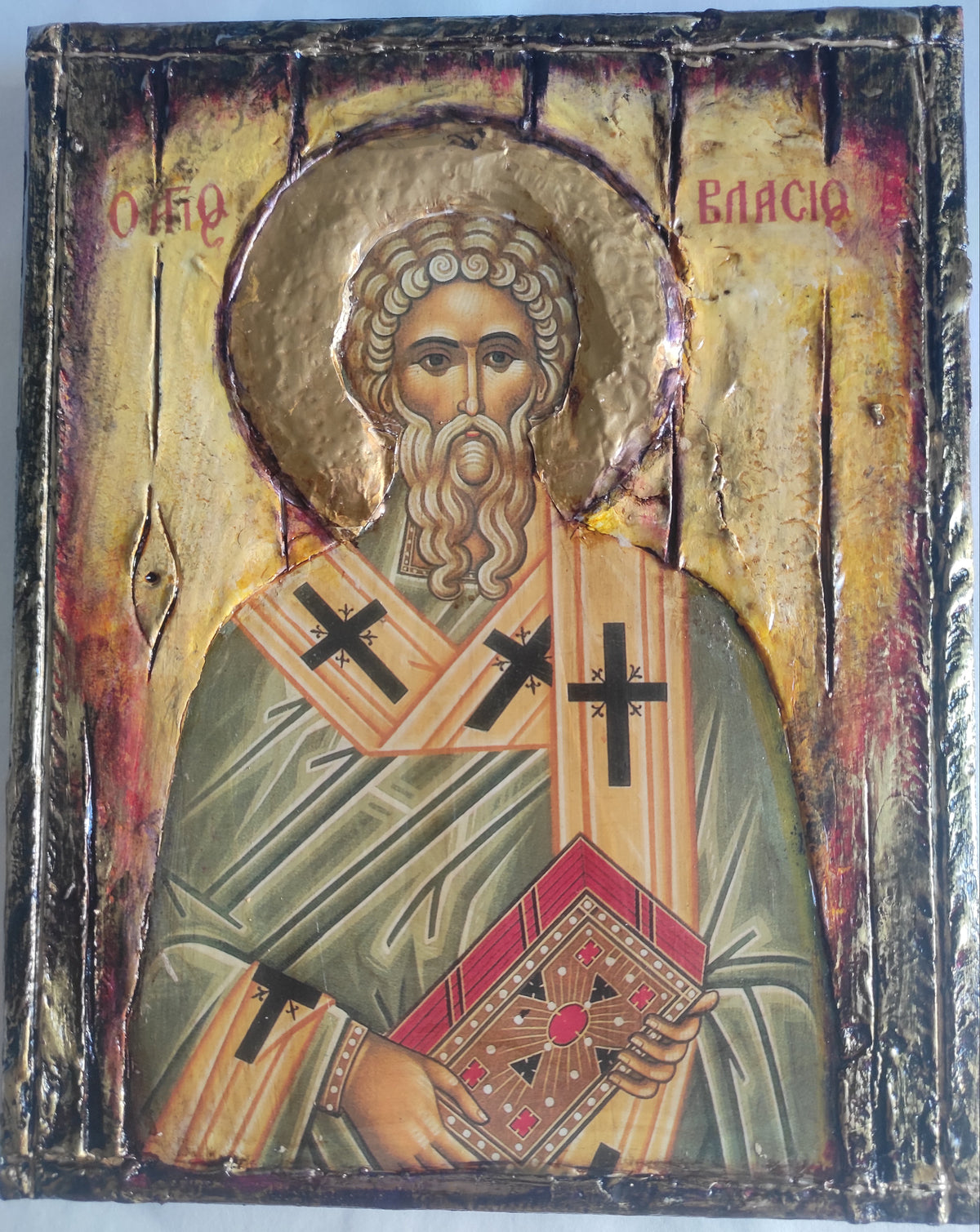 A rectangular religious icon design on a textured wooden panel, depicting Saint Basil with a gold halo, green and beige robes, and a red patterned book, photographed against a light gray background.