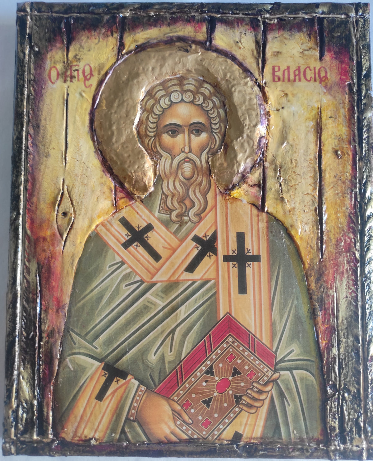 An antique religious icon design of Saint Vlasis, featuring a gold-leaf background, a halo. The saint is depicted wearing green and gold robes with black crosses, holding a red and gold book.
