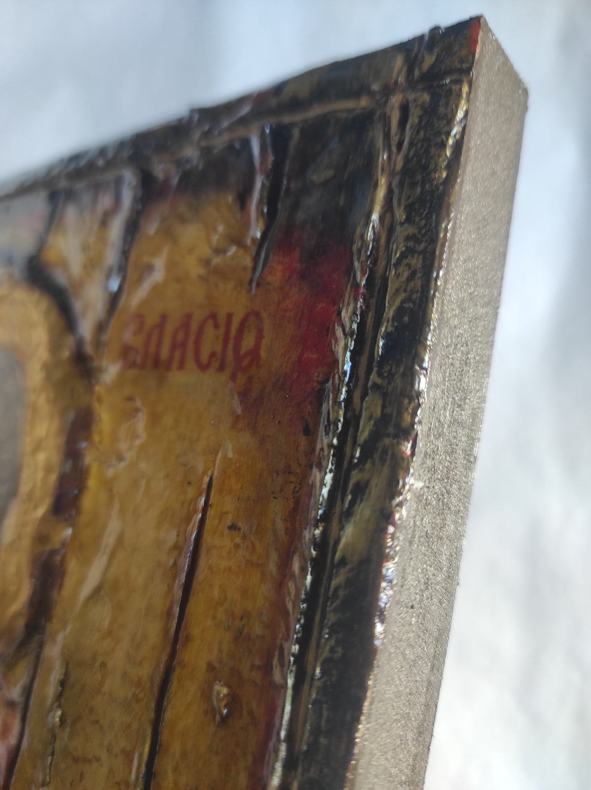 Close-up of a textured religious icon or plaque showing a gold-toned surface with red designed lettering, dark metallic detailing, and a light gray stone or plaster edge against a white background.