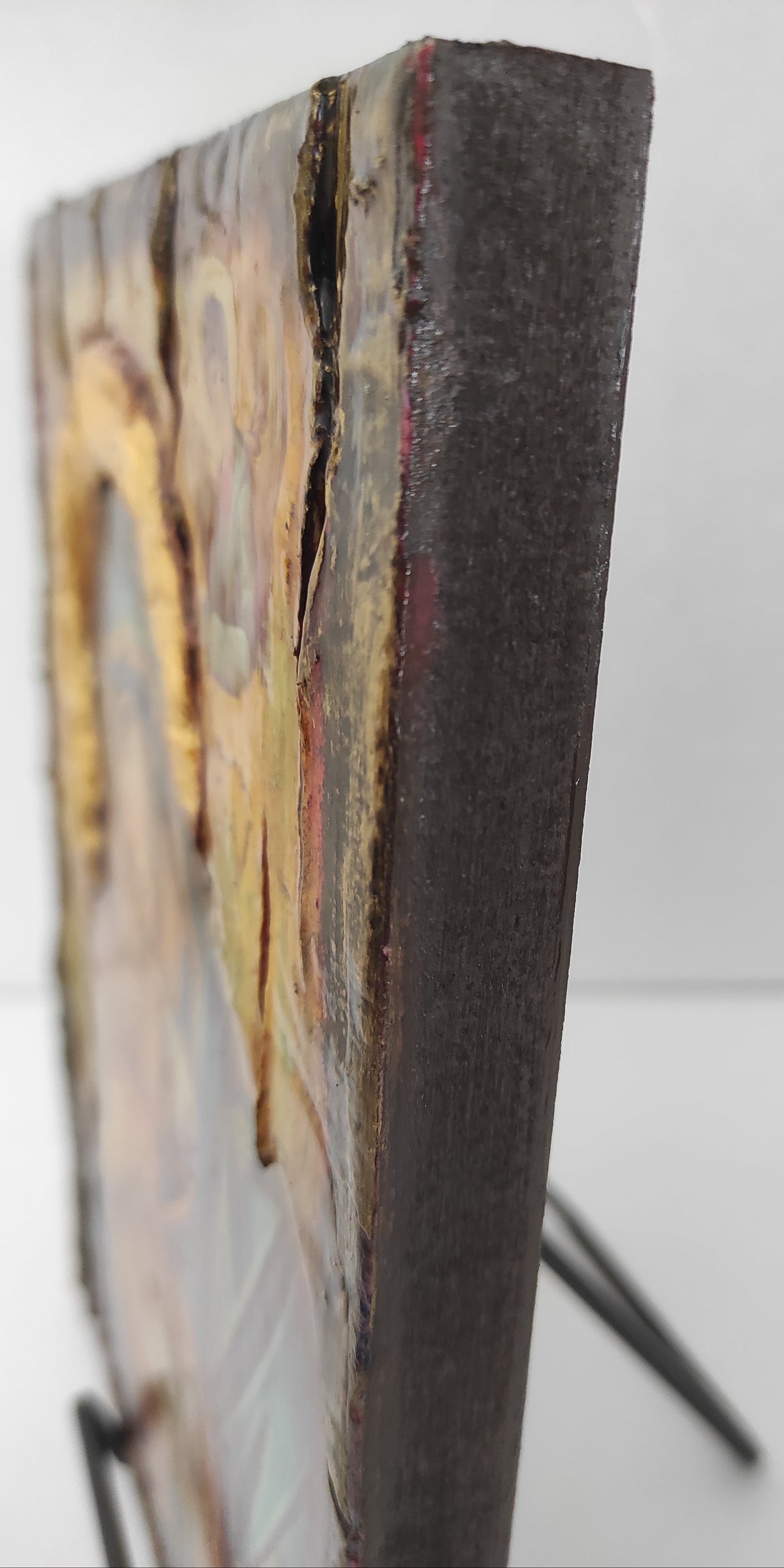 A close-up of a rectangular wall art piece showing a dark wooden edge and designed  surface with gold, beige, and brown tones, mounted on a black wire display stand against a white background.