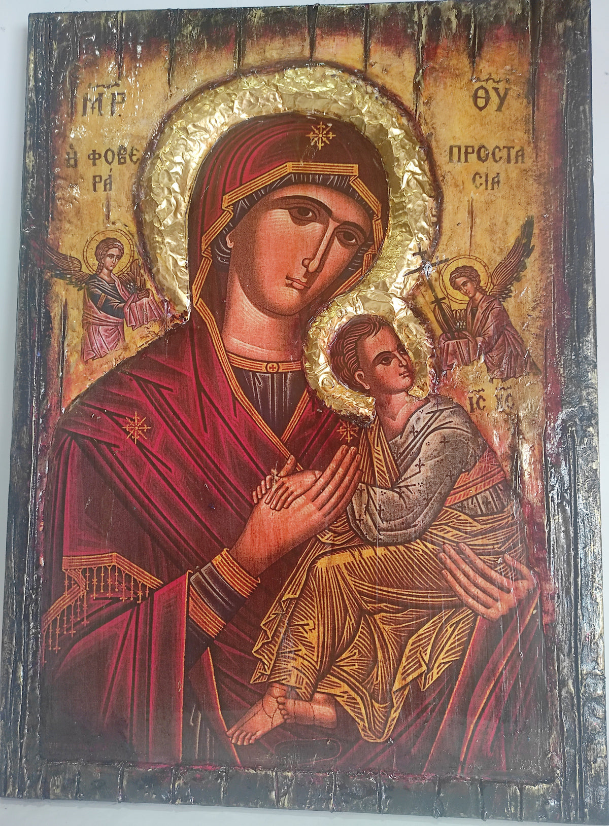 A traditional Eastern Orthodox religious icon design of the Virgin Mary holding the Baby Jesus, featuring a gold leaf halo and deep red robes on a wooden panel.
